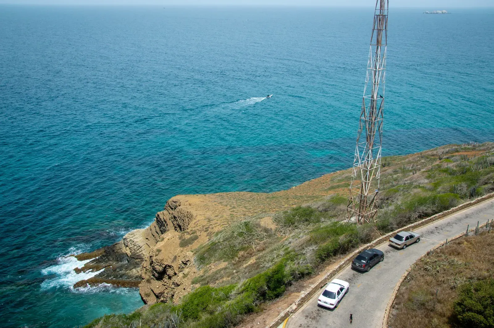 Aerial view of cars on a coastal road alongside a towering structure by the blue sea.