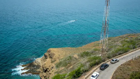 Aerial view of cars on a coastal road alongside a towering structure by the blue sea.