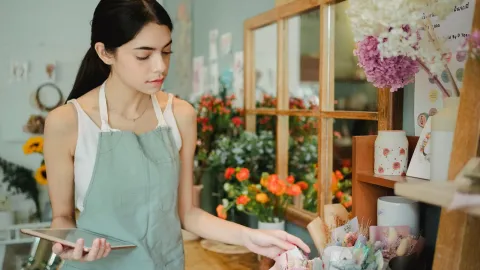 Concentrated female florist with dark hair standing in floral shop with tablet near flowers in wrapper while checking or
