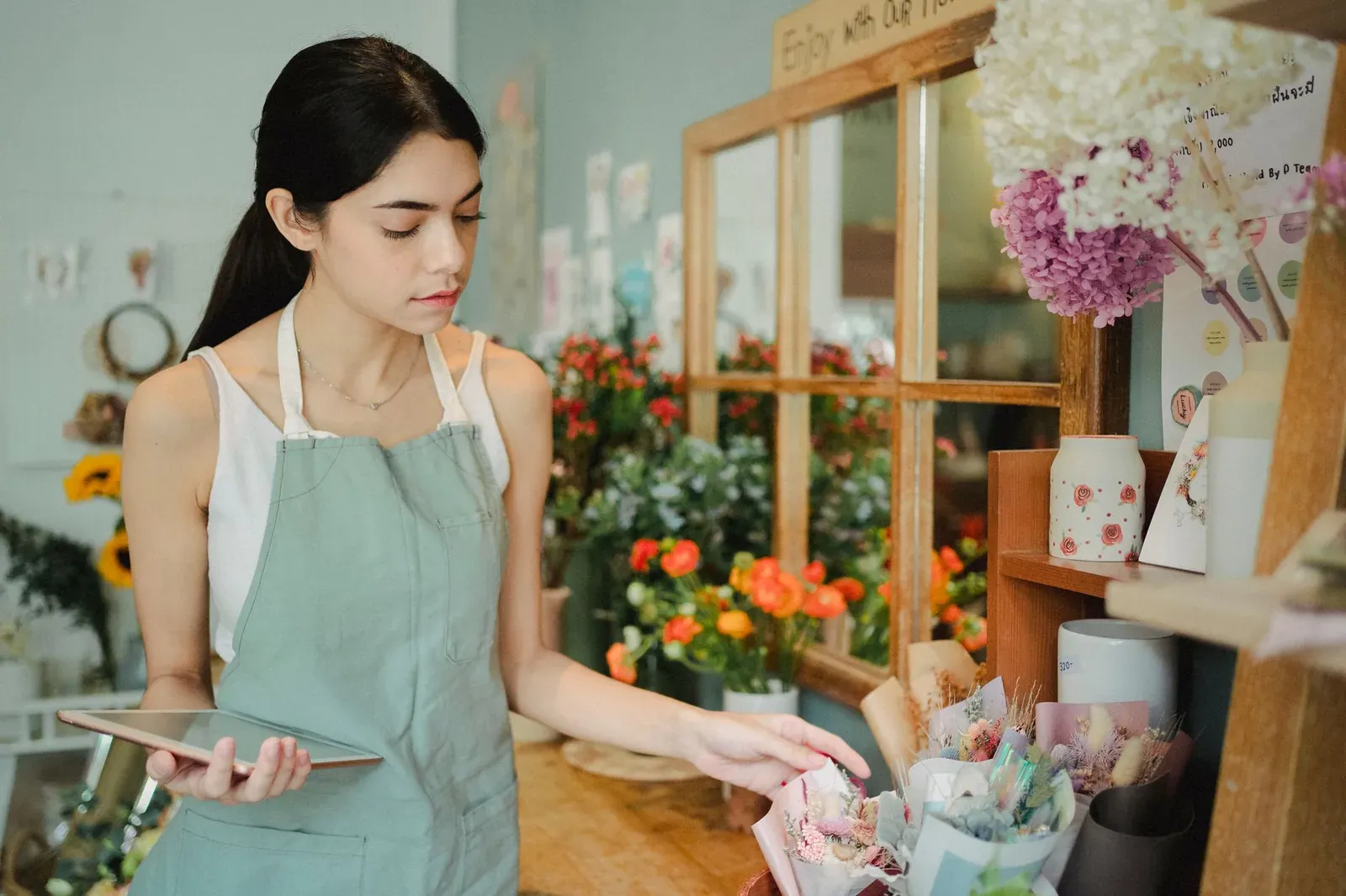 Concentrated female florist with dark hair standing in floral shop with tablet near flowers in wrapper while checking or