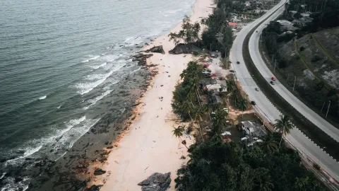Aerial view of a picturesque coastline with winding road, palm trees, and crashing waves.