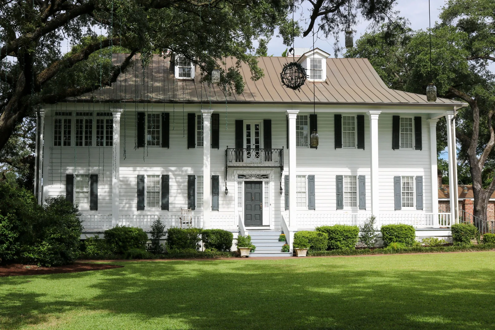 A classic white Lowcountry home with columns, a metal roof, and black shutters beneath live oak trees