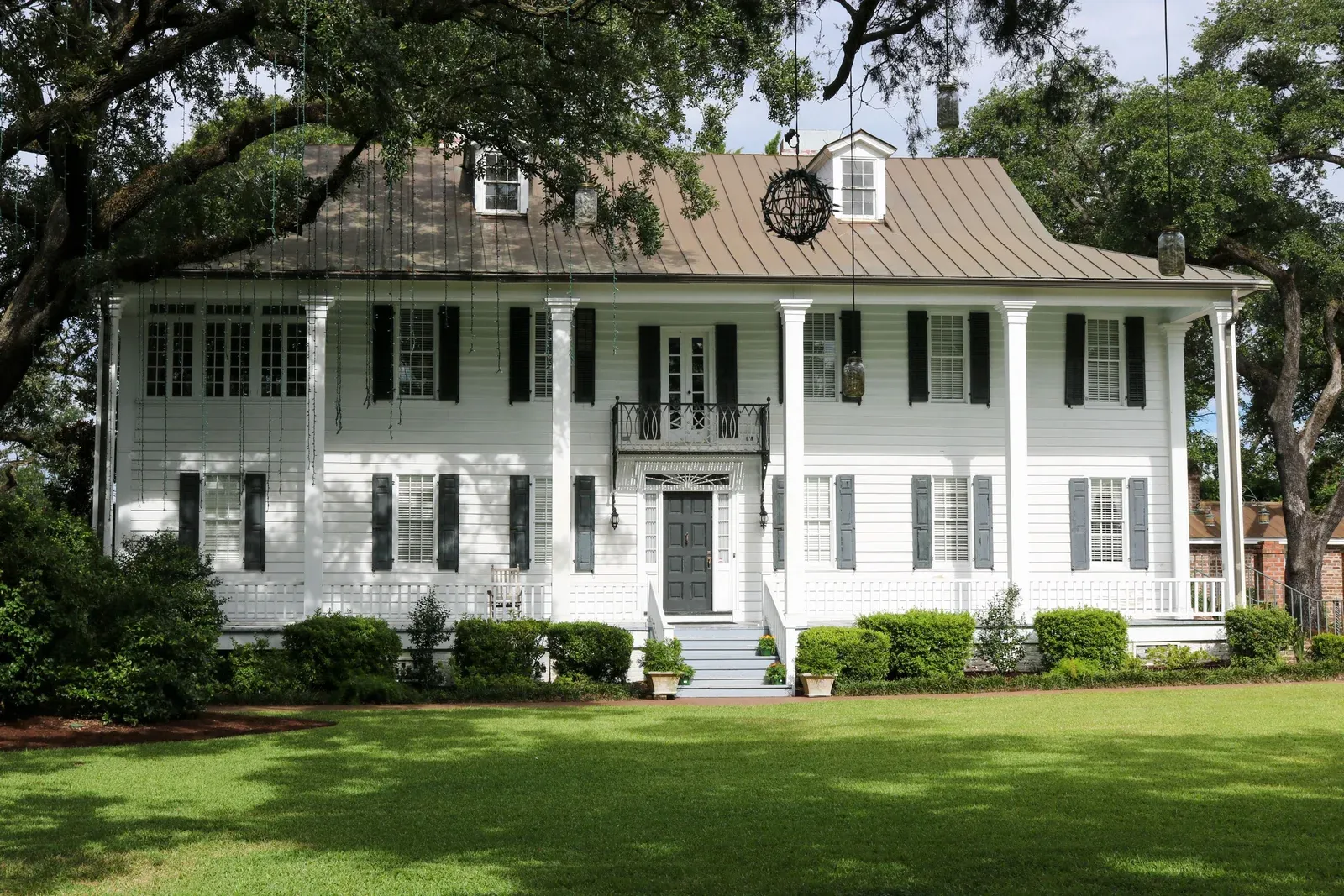A classic white Lowcountry home with columns, a metal roof, and black shutters beneath live oak trees