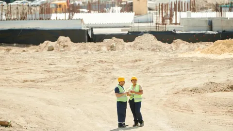 Two workers discussing plans on a sandy construction site, wearing safety gear.