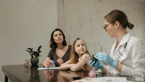 A doctor in a white coat consults a young girl and her mother about health in an indoor setting.