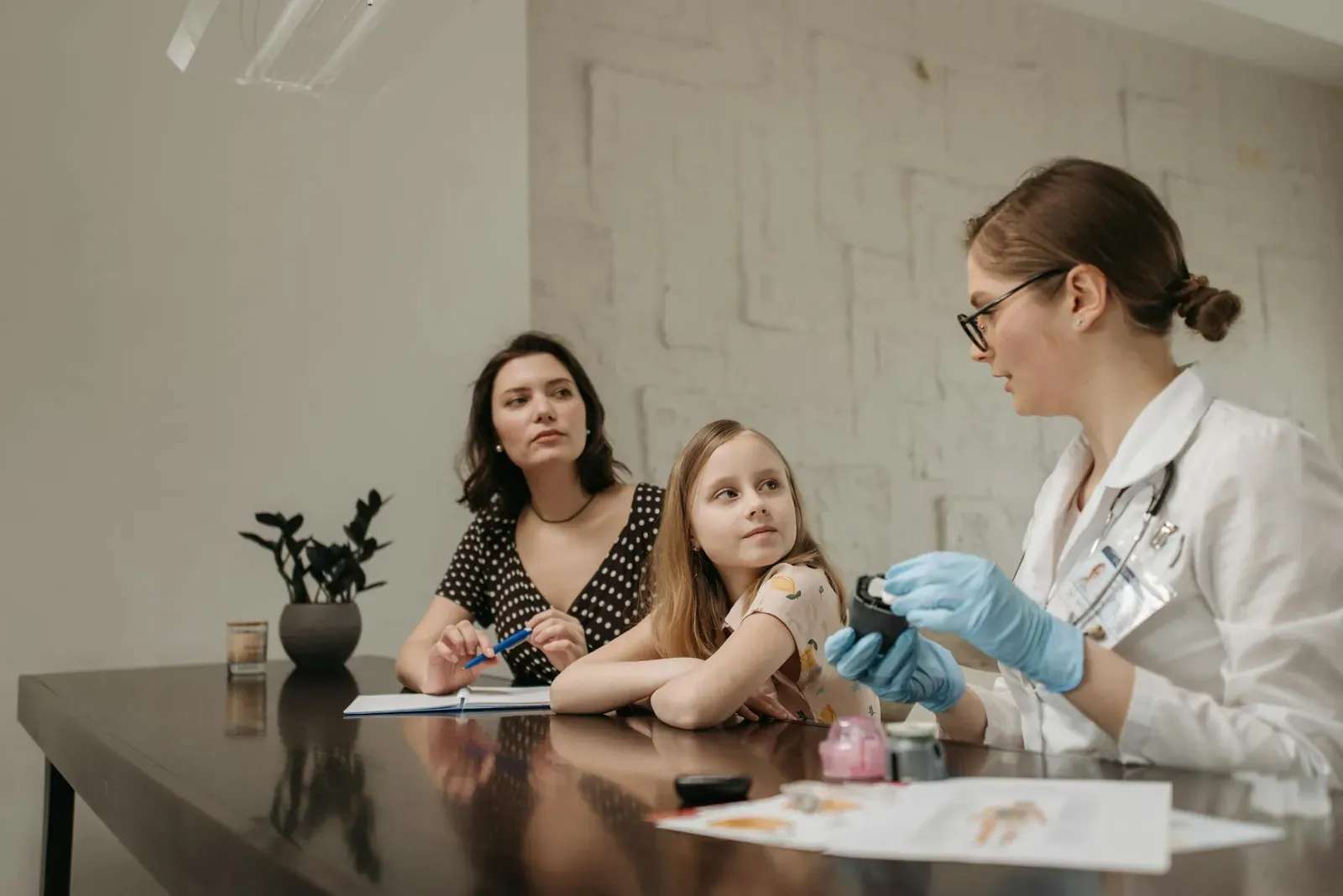 A doctor in a white coat consults a young girl and her mother about health in an indoor setting.
