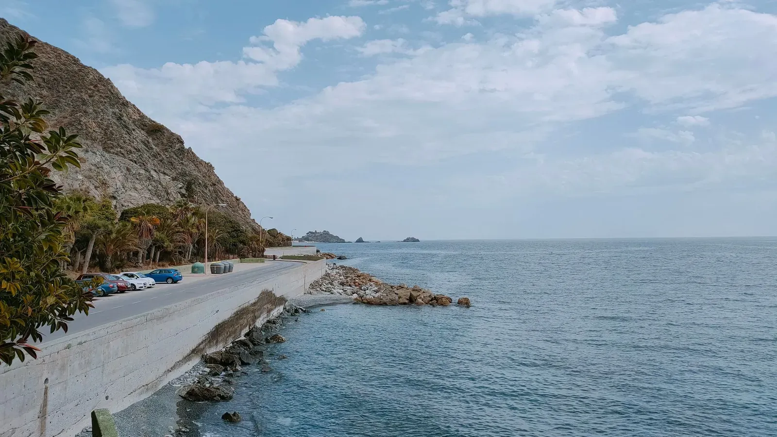 Serene view of a coastal road next to a calm ocean and rocky seashore under a partly cloudy sky.