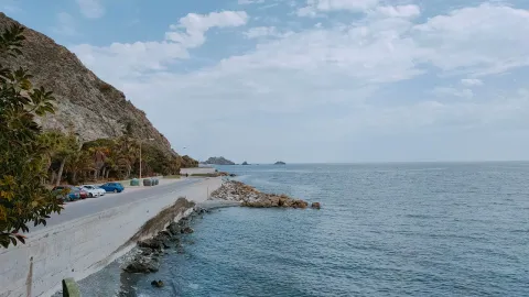 Serene view of a coastal road next to a calm ocean and rocky seashore under a partly cloudy sky.