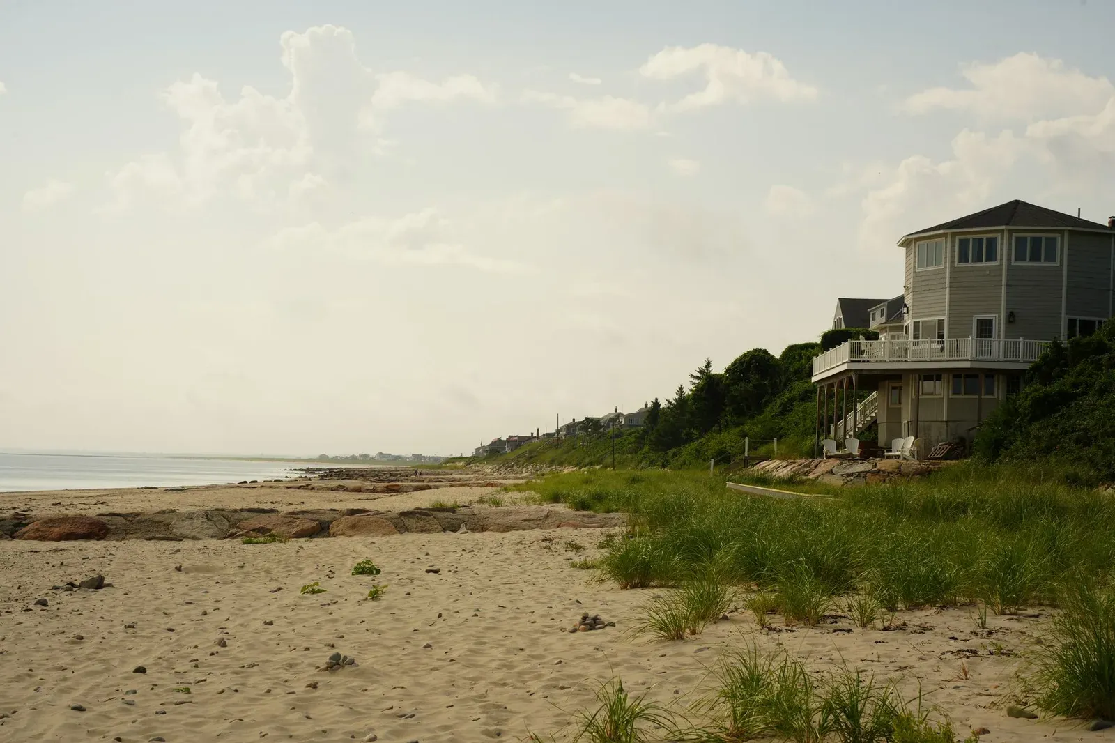 A raised coastal beach house behind sandy dunes and sea grass along the shoreline.