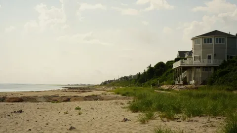 A raised coastal beach house behind sandy dunes and sea grass along the shoreline.