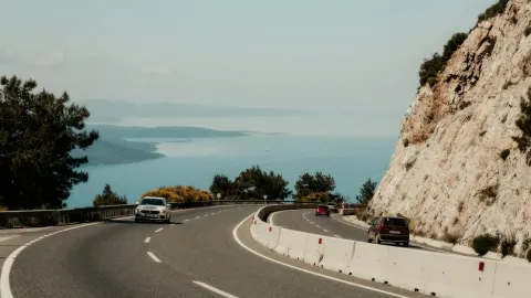 A winding coastal road with cars, overlooking a serene sea and rocky cliffs.