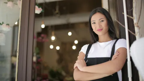 From below of young confident Asian female standing with crossed arms at entrance of floral shop and smiling at camera