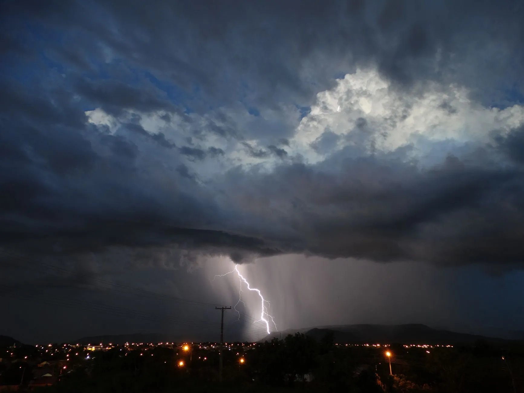 Glowing lightning striking in dark sky with stormy clouds over city with residential buildings with shining lights at night time