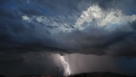Glowing lightning striking in dark sky with stormy clouds over city with residential buildings with shining lights at night time
