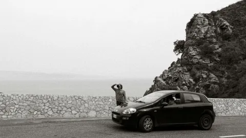 Black car with a person standing by a rocky mountain roadside in grayscale.