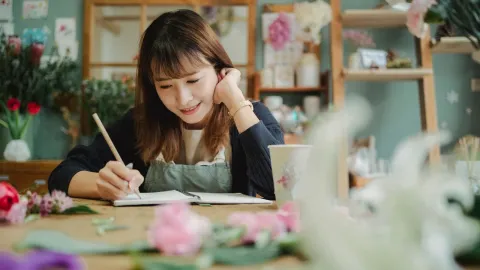 Smiling young female florist in apron writing notes while working in cozy flower shop