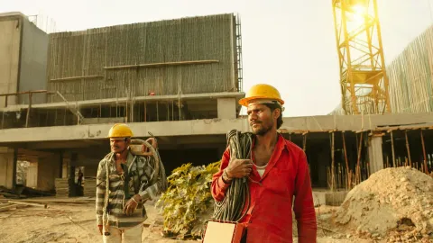 Two construction workers wearing helmets at a site in India with a crane and building.